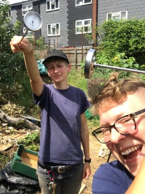 2 queers with matching t-shirts wield long metal thermometers in a garden in a residential area, looking pleased with themselves.