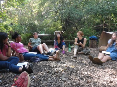 7 people are sitting in a circle on the ground, surrounded by trees, one person is reading from a book whilst the others listen