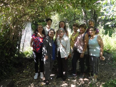 a multigenerational group (mostly elders) stand together in dappled light in a garden smiling.