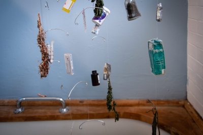 side view of mobile, against blue bathroom wall. Hanging objects include: dried herbs, pill packets, syringe, laughing gas cannister, packagings in different languages, a cigarette box, a glass bottle containing soil 
