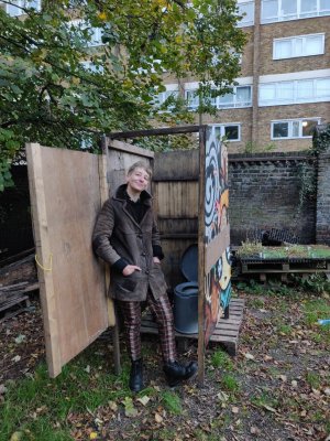 Hari wearing brown coat and chequered trousers stands in toilet doorway smiling, the grey plastic toilet is visible inside