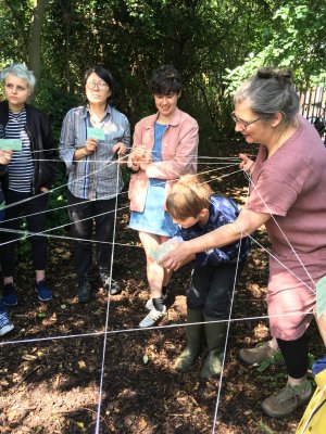 adults and a child stand around connected by string, which looks like a giant cats cradle. They are smiling / looking thoughtful