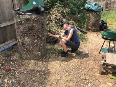 person kneels in front of large round thermal compost pile, inserting a thermometer and reading the temperature. A 2nd thermal pile / cylinder is in the background