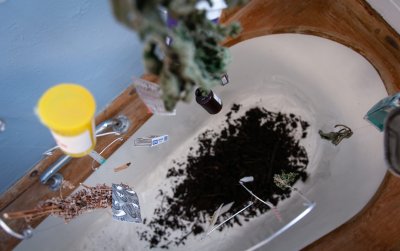 birds eye view of mobile hanging over bath containing compost, the objects in the mobile include:  Objects include: dried herbs, pill packets, syringe, a cigarette butt